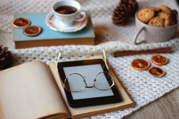 Cup of tea, books, tablet, bowl of cookies, various spices, pine cones and lit candles. Hygge at home. Selective focus.