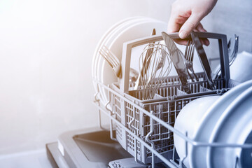 Close-up of female hand holding full cutlery basket with clean knife, fork, whisk. Loading to,...