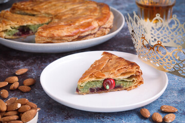 A cut piece of traditional French galette des rois or King cake, fève inside and almond with glasses of apple cider. Cake made with puff pastry and creamy almond filling. With crown in background 