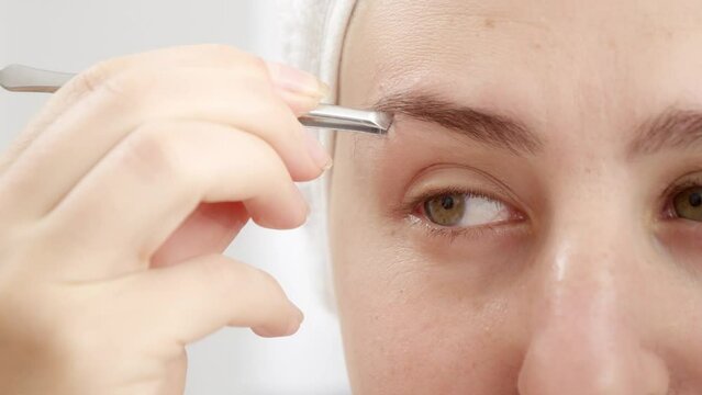 Macro shot of woman plucking eyebrows with small cosmetic tweezers at home. Concept of beautiful female, makeup at home, skin care and domestic beauty industry.