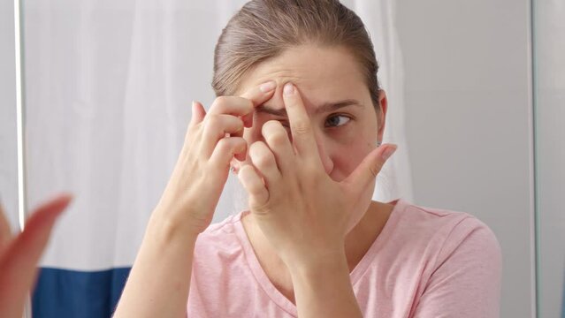 Beautiful young woman looking for pimples and squeezing them in bathroom. Concept of female aging, skin issues, facial treatment and care