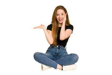 Obraz premium Young redhead woman sitting on the floor cut out isolated holds copy space on a palm, keep hand over cheek. Amazed and delighted.