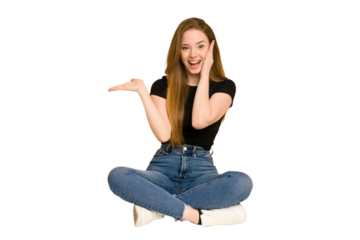 Young redhead woman sitting on the floor cut out isolated holds copy space on a palm, keep hand over cheek. Amazed and delighted.