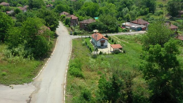 Aerial view of a church with bell tower in the heart of a small balkan village with a white house and a brown roof, a cloudy sky and a forest on the horizon.