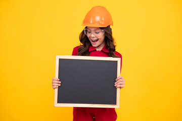 Child girl builder in hard hat helmet. Teenage girl worker hold blackboard isolated on yellow background. Kids renovation concept. Copy space, mock up.
