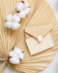 Blank card and envelope on dried palm leaf with cotton flowers top view, wedding mockup