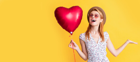 happy girl in straw hat and sunglasses hold love heart balloon on yellow background. Woman isolated...