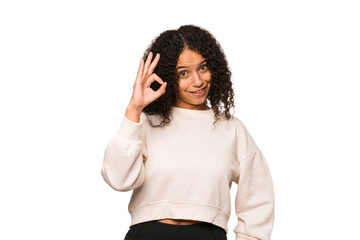 Young african american curly woman isolated winks an eye and holds an okay gesture with hand.