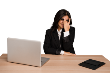 Young indian woman in a table with a laptop and tablet isolated blink at the camera through fingers, embarrassed covering face.