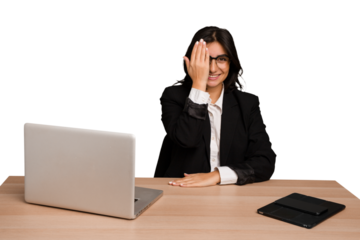 Young indian woman in a table with a laptop and tablet isolated having fun covering half of face with palm.