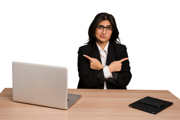 Young indian woman in a table with a laptop and tablet isolated points sideways, is trying to choose between two options.