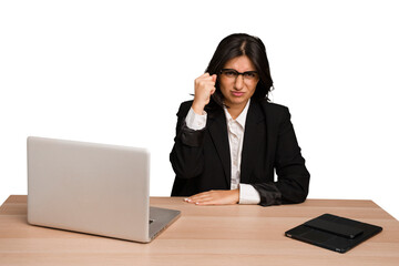 Young indian woman in a table with a laptop and tablet isolated showing fist to camera, aggressive facial expression.