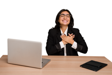 Young indian woman in a table with a laptop and tablet isolated laughing keeping hands on heart, concept of happiness.