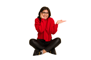 Young indian woman sitting on the floor cut out isolated holds copy space on a palm, keep hand over cheek. Amazed and delighted.