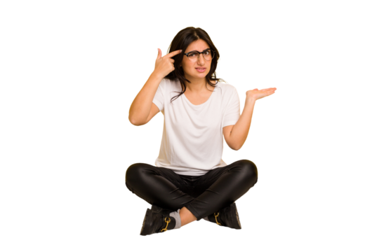 Young indian woman sitting on the floor cut out isolated holding and showing a product on hand.