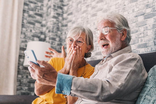 Happy older couple sit on couch staring at cellphone screen gesturing looking overjoyed, scream with joy, read fantastic news, get great commercial offer, pension raise. Success, achievement concept.