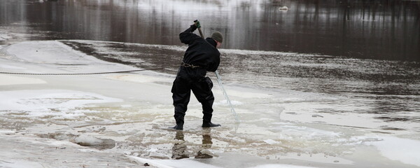 Lifeguard diver saws ice on the river with a big hand saw at winter day