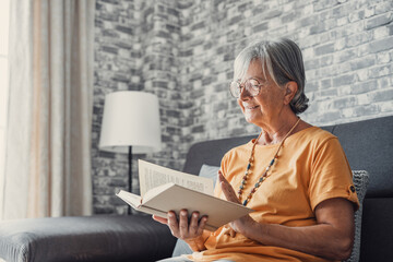 Happy older blonde woman sitting on comfortable sofa in living room, reading interesting book. Smiling elder retired grandmother enjoying leisure weekend time with favorite novel alone at home..