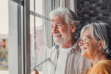 Happy bonding loving middle aged senior retired couple standing near window, looking in distance,...