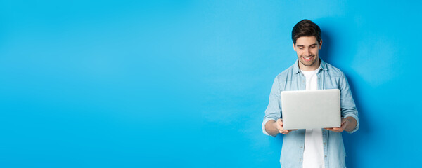 Handsome man working on laptop, smiling and looking at screen satisfied, standing against blue...