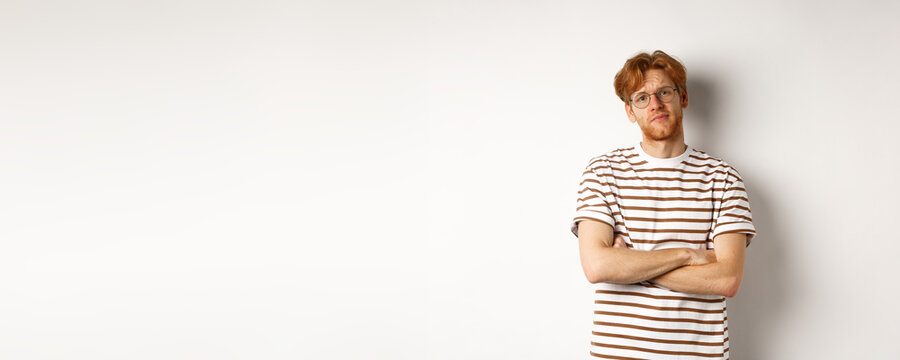 Teenage Redhead Guy In Glasses Cross Arms On Chest, Looking Skeptical And Unamused At Camera, Standing Over White Background