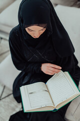 Young traditional muslim woman reading Quran on the sofa before iftar dinner during a ramadan feast at home