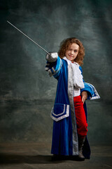 Emotional kid, little girl wearing costume of prince, musketeer and royal person posing over dark...