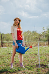 Young cute blondie girl  in hat, beige shirt, denim shorts, yellow rubber gloves  smiling and watering in the garden the fruit tree with blue watering can