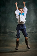 Naklejka na ściany i meble Emotional kid, little girl wearing costume of prince, musketeer and royal person posing over dark vintage style background. Fashion, theater, beauty, emotions concept
