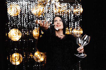 Happy woman with brunette long hair in trendy evening fur coat holding glass and confetti on shine wall in studio