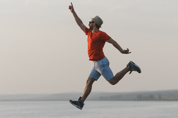 Young hipster man jumping up in the air at picturesque view of Italian lake  in hat, red t-shirt, blue shorts 