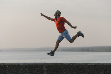 Young hipster man jumping up in the air at picturesque view of Italian lake  in hat, red t-shirt, blue shorts 