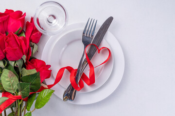 Fototapeta na wymiar Valentine day table setting on white background. Romantic diner table setting for with red roses, heart shaped bow, plate, wine glass and cutlery top view copy space. Valentines menu background