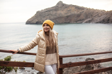 Fototapeta premium A beautiful woman walks on the beach in cold weather, meditates to the sound of the waves. The girl dressed sweater, white jeans and yellow hat