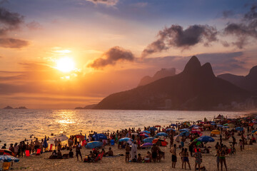 Beachgoers in Rio de Janeiro, Brazil play on the sand and in the water of the Atlantic Ocean as the sun sets over the mountains above Ipanema Beach.
