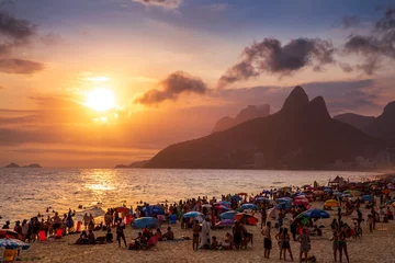 Papier Peint photo autocollant Copacabana, Rio de Janeiro, Brésil Beachgoers in Rio de Janeiro, Brazil play on the sand and in the water of the Atlantic Ocean as the sun sets over the mountains above Ipanema Beach.