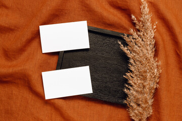 Two blank paper sheet cards on dark wooden tray on linen brown cloth with pampas grass, flat lay, top view. Minimal business card with copy space, invitation, aesthetic branding