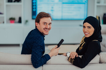 Young muslim couple woman wearing islamic hijab clothes sitting on sofa watching TV together during the month of Ramadan at modern home