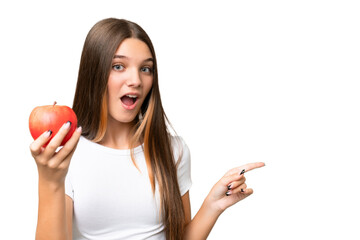 Teenager caucasian girl holding an apple over isolated background surprised and pointing finger to the side