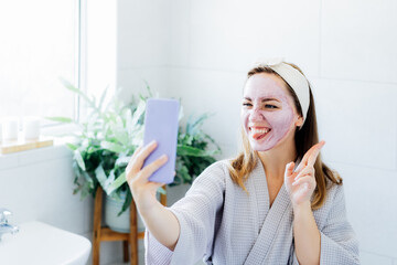 Smiling woman with facial pink clay mask taking selfie with mobile phone at home bath enjoying relaxation and spa beauty treatment. Teen Beauty blogger creating content. Selective focus, copy space.