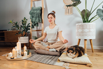 Young healthy woman practicing meditation at home, sitting in lotus position on floor cushion with relaxed cat pet in modern Scandi interior home. Meditating, relaxed, eyes closed. Selective focus - Powered by Adobe