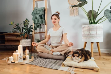  Young healthy woman practicing meditation at home, sitting in lotus position on floor cushion with relaxed cat pet in modern Scandi interior home. Meditating, relaxed, eyes closed. Selective focus © okrasiuk