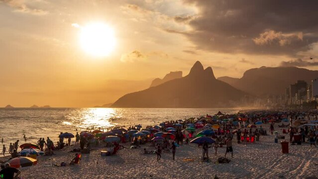 A timelapse of beachgoers in Rio playing on the sand and in the water of the Atlantic Ocean as the sun sets over the mountains above Ipanema Beach.