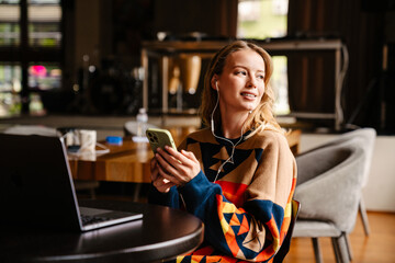 Young woman holding smartphone and looking out of window while working on laptop in cafe