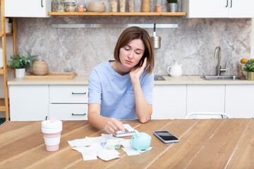 Unhappy and depressed woman sitting in the kitchen at the table, holding piggy bank and looking at a lot of bills and checks. Taxes and debts, family budget and spending concept