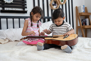 Fototapeta na wymiar Cute small boy and his sister sitting try to play guitar on bed in the bedroom