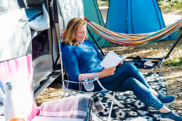 Adult woman relaxing, sitting in the travel chair and reading book on the background of camper car vehicle. Female living on camper car and travel the world. Caravan car Vacation. Adventure Is Ageless