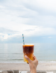 A glass of fresh iced orange americano decorated with sliced ​​orange holding by hand on the beach view, seascape, vertical style. Cold black coffee with orange juice, mixing mocktail drink menu.
