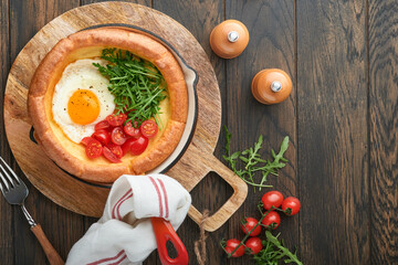 Dutch baby pancake. Fresh homemade Dutch Baby pancake with fried egg, tomato and green arugula in reed cast-iron pan on old wooden table. Flat lay, close up.
