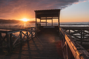 Bird hide on a wooden pier at sunset on Playa America beach. Sun star over the mountains on the coastline. Nigran, Pontevedra, Galicia, Spain.